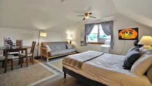 A cozy room with a bed, a sofa, a desk, and a dining table. There is a ceiling fan, a window with curtains, and a sign on the wall.