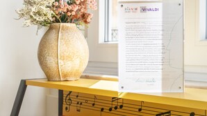 Um piano com partituras, um vaso com flores e uma janela ao fundo.