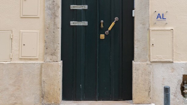 Uma porta dupla verde escura com maçaneta de latão e fechadura, uma pequena janela acima dela, e uma placa com o número 16.