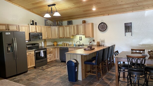 A modern kitchen with wooden cabinets, a built-in oven, a refrigerator, and a microwave. There is a dining area with a bar stool and chairs. - Cozy Cabin get away - walking distant to beach (Crofton)