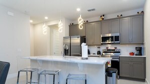 A modern kitchen with a central island, stainless steel appliances, and clear bar stools. - Exclusive at Reunion Golf View Resort Style 764 (Celebration)