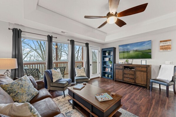 A spacious living room with a ceiling fan, large windows, a bookshelf, and a painting on the wall.