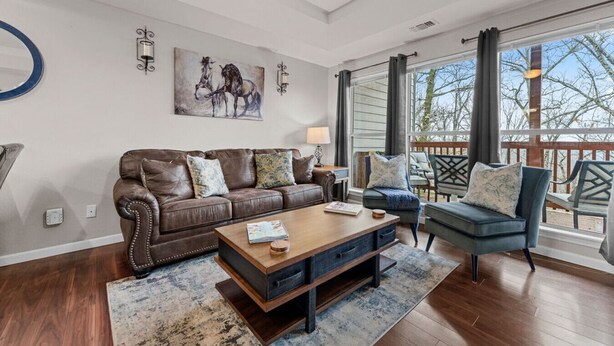 A living room with a brown leather couch, a wooden coffee table, and a blue armchair. There is a painting on the wall and a window with curtains. - Spacious remodel 5 mn-SDC, 10 mn-Lake, 2BR 2.5BA (Branson West)