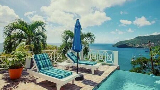 A poolside area with lounge chairs and a blue umbrella, overlooking a body of water and a hilly landscape.