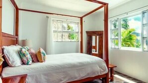 A four-poster bed with a white bedspread, a wooden headboard, and multiple pillows. There is a window with a view of greenery outside.