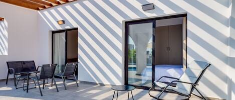 Un patio moderno al aire libre con una pérgola de madera, un ventanal que da a una habitación y una zona de estar con sillas y una mesa.