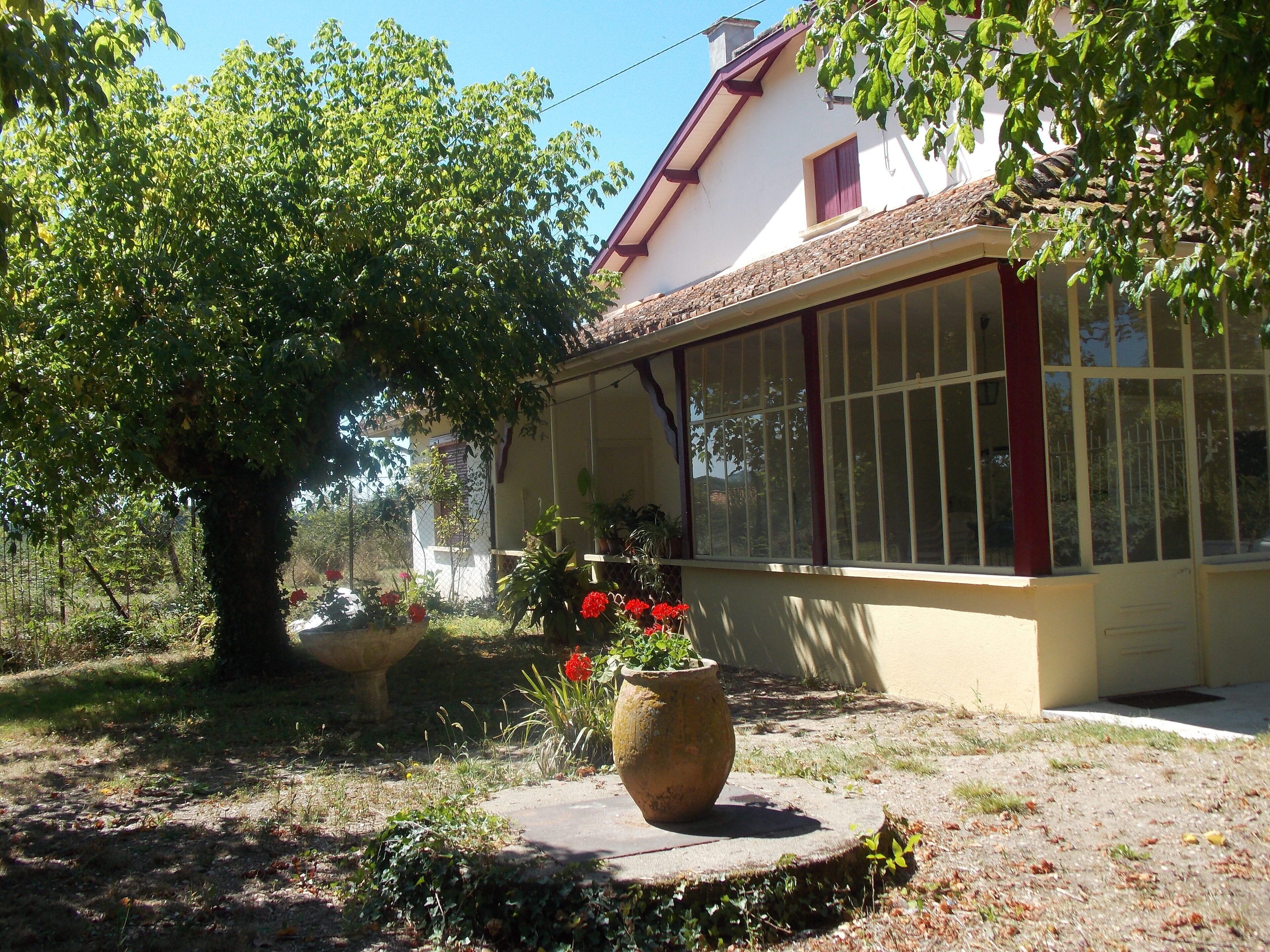 Une maison avec une grande fenêtre, un jardin avec un arbre et des plantes en pot, et un banc en pierre.