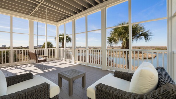 A covered outdoor deck with wicker furniture, a wooden table, and a view of the water and palm trees.