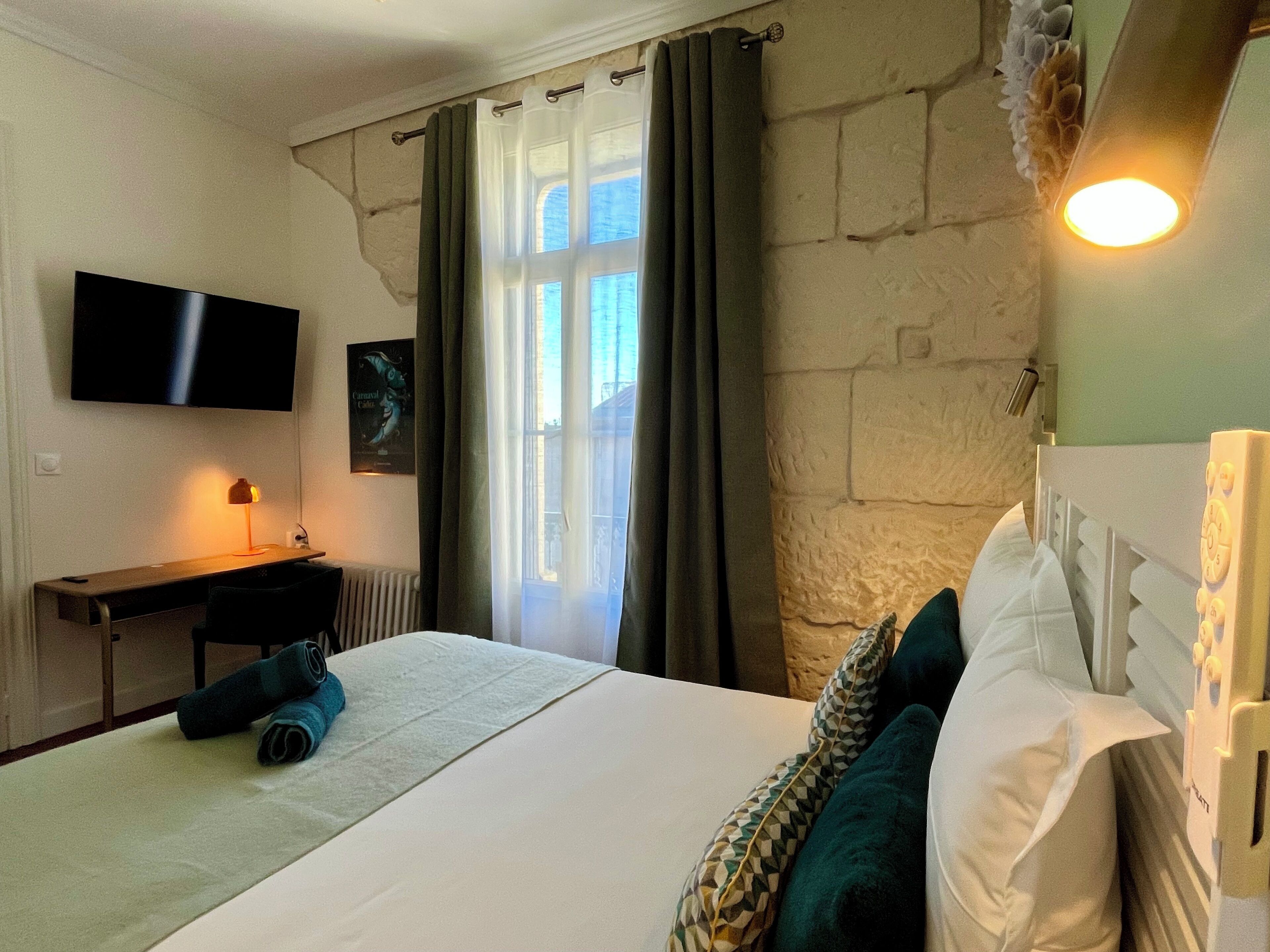Habitación de hotel con cama, escritorio, televisor, ventana con cortinas y lámpara de pared.