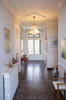 Un pasillo con baldosas de diseño, un radiador blanco, una mesa de madera con objetos decorativos, una araña y una puerta con paneles de vidrio.