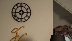 A wall clock with Roman numerals, a decorative letter 'GE' sign, a desk with a lamp, and a mirror. - Harakop Otel (Ayvalik)