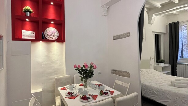 Una habitación de hotel moderna con comedor que cuenta con una mesa blanca dispuesta para cuatro personas, rodeada de sillas blancas. Se observa un aparador rojo con objetos decorativos y un reloj. La habitación tiene piso de madera y un ventanal con cortinas.