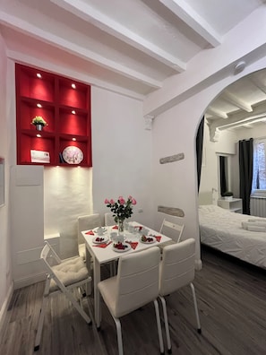 Quarto de hotel moderno com área de refeições, apresentando uma mesa branca posta para quatro pessoas, rodeada por cadeiras brancas. Uma estante vermelha com objetos decorativos e um relógio é visível. O quarto tem piso de madeira e uma grande janela com cortinas.