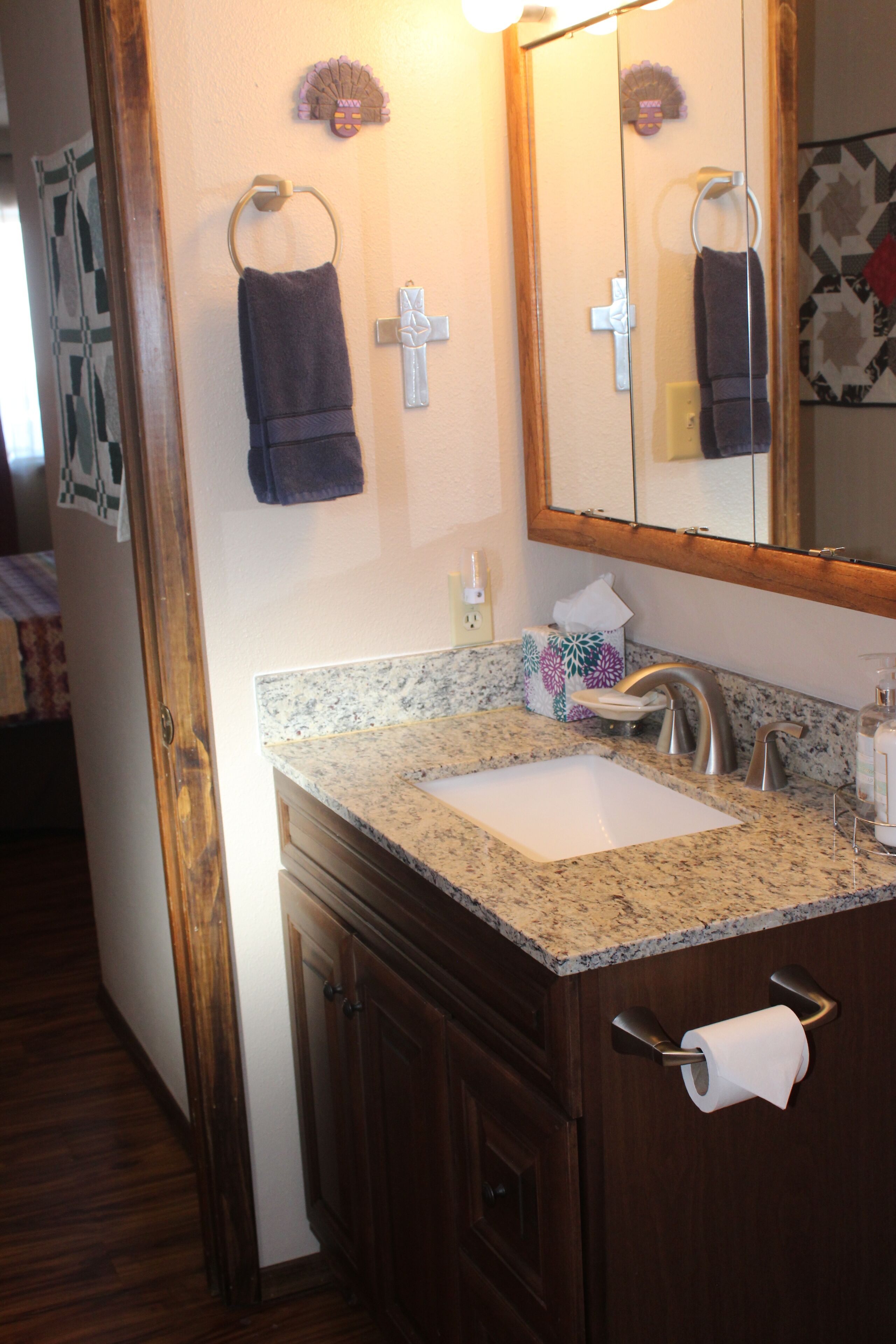 Une salle de bain avec un lavabo surmonté d’un plan de travail en granit, des meubles en bois, un porte-serviettes et un miroir orné d’une croix.