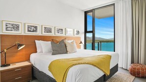 A modern hotel room with a large bed, a nightstand, a lamp, and a view of the sea through a large window.