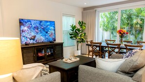 Un salon avec un téléviseur, une table basse en bois foncé, des canapés gris, une table à manger avec des chaises, une plante et une fenêtre avec des rideaux.