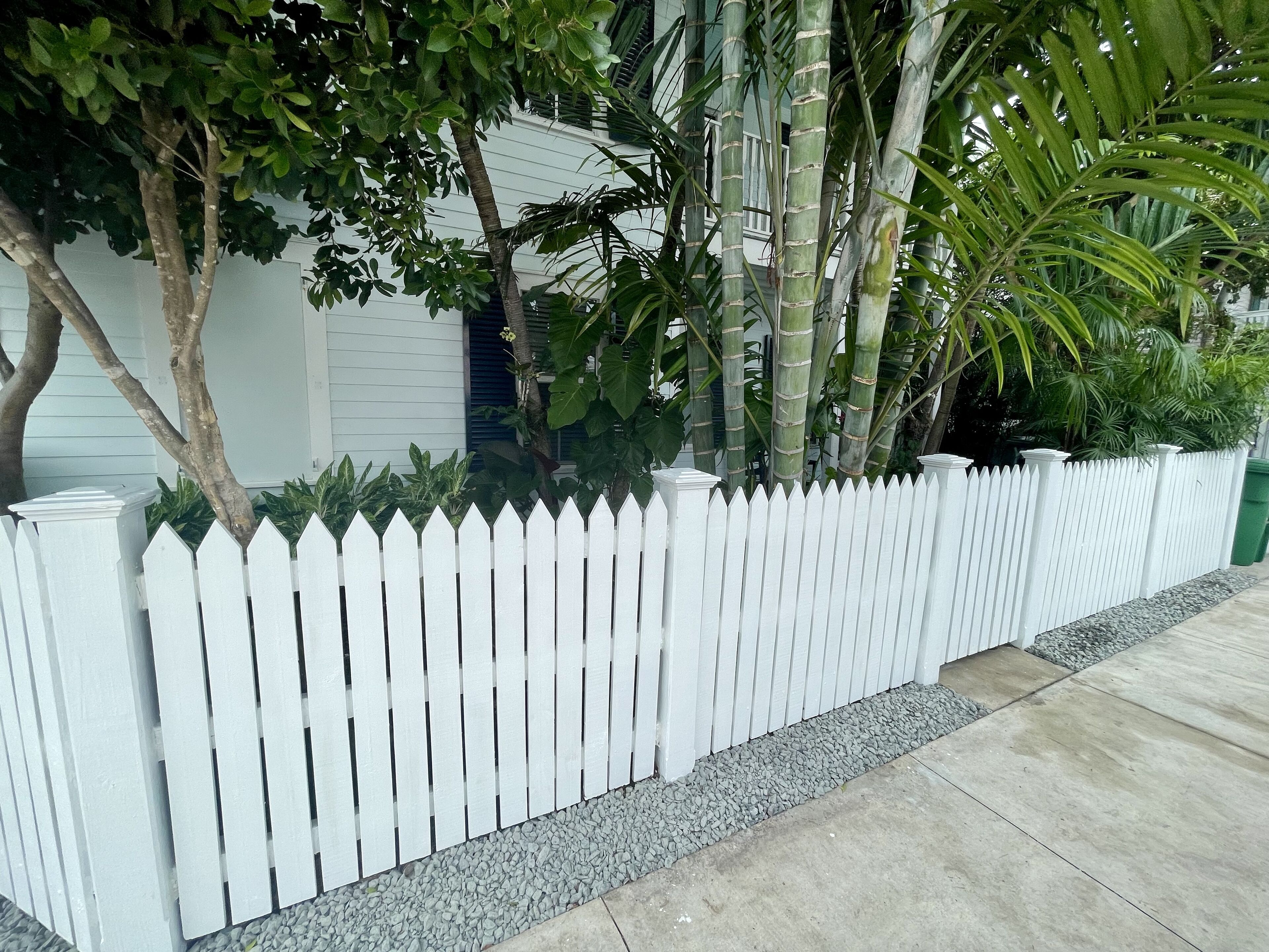 A white picket fence with a gravel border, a house with white siding, and various green plants and trees in the background.
