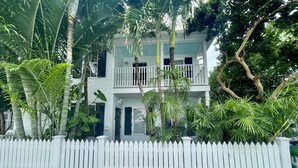 Une maison à deux étages, avec une clôture en bois peinte en blanc, entourée d’une végétation luxuriante et de palmiers.