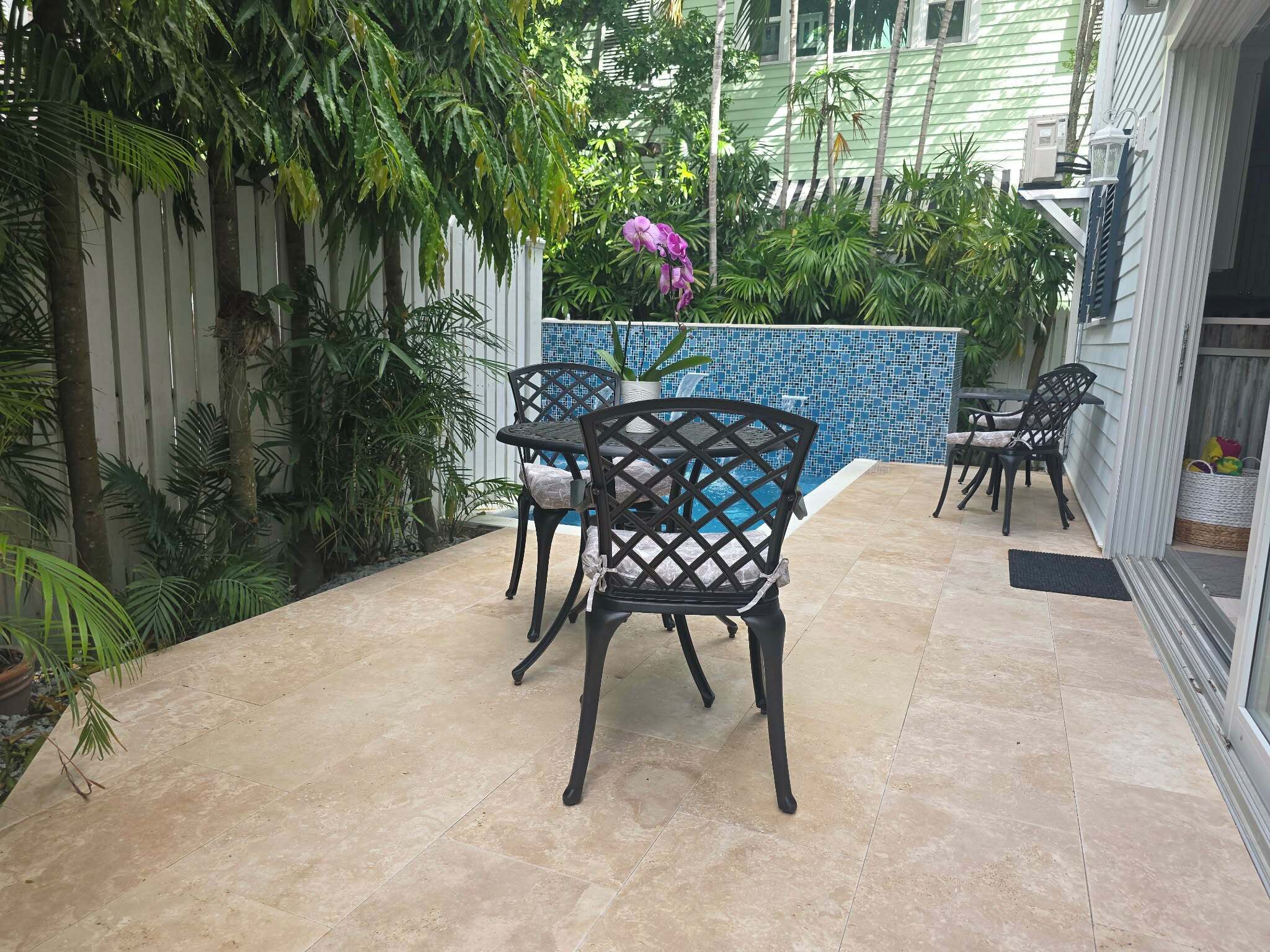 An outdoor patio with a pool, surrounded by lush greenery and a white fence.