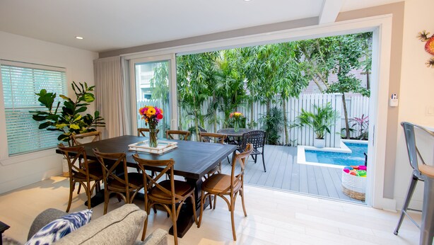 Un coin repas avec une table et des chaises, un canapé et une vue sur un jardin avec une piscine et des plantes.