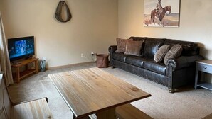 Een woonkamer met een houten salontafel, een leren bank, een hangend schilderij, een plafondventilator en een televisie op een standaard.