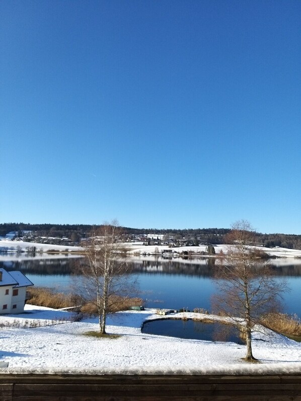 A serene lakeside view with a small house, snow-covered ground, and clear blue sky. - CHALET WITH SPA AND LAKE VIEW (Montperreux)
