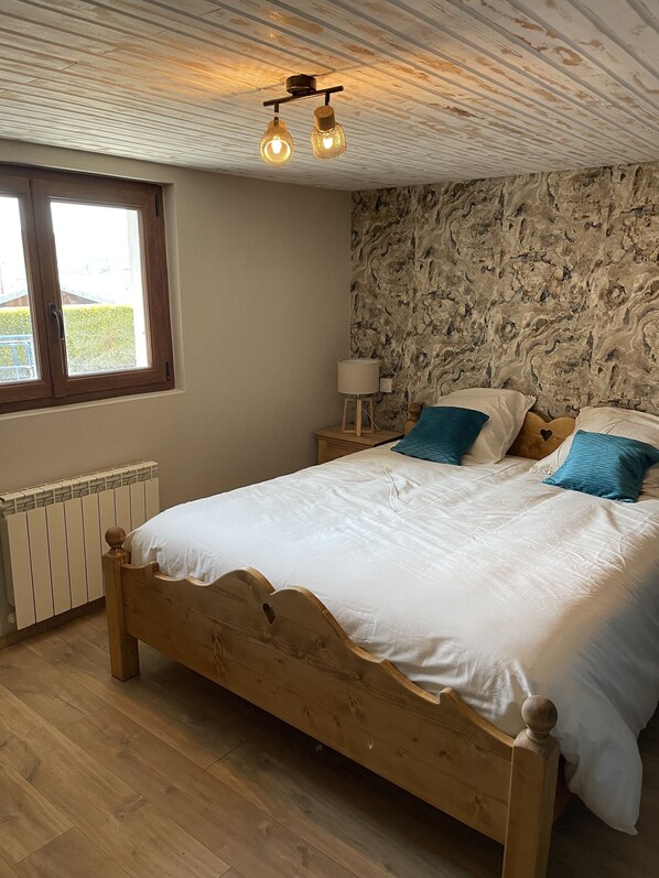 A bedroom with a wooden bed, a nightstand with a lamp, a stone-patterned wall, and a window with a view of greenery. - CHALET WITH SPA AND LAKE VIEW (Montperreux)