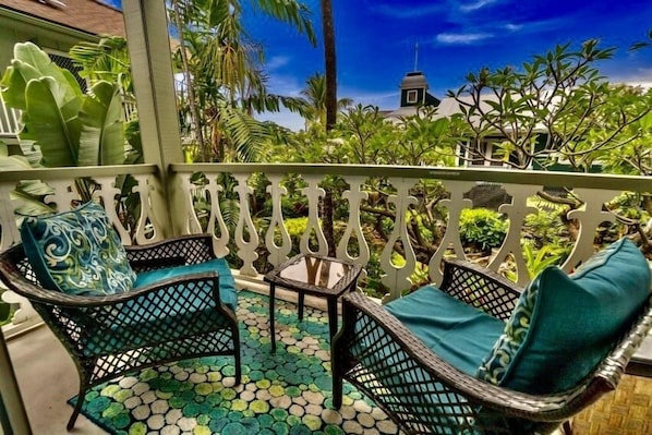 A balcony with wicker furniture, a small table, and a view of greenery and a building in the background.
