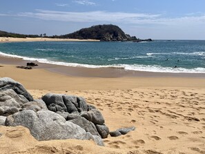 A beach with large rocks in the foreground, a sandy shore, and a distant hill.