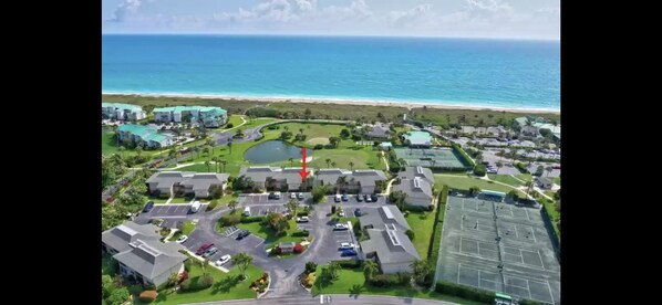 An aerial view of a coastal residential area with a golf course, a large parking lot, and a beach in the background.
