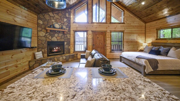 Interior de una cabaña de madera con chimenea de piedra, una cama grande, un sofá y un comedor con una mesa de granito.