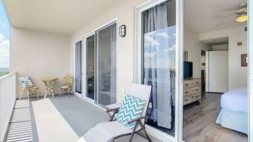 Een balkon met rieten stoelen en een tafel, een schuifdeur die leidt naar een slaapkamer met een bed en een dressoir.