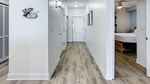 Ein Flur mit Holzboden, weißen Wänden und einer Tür am Ende, die zu einem Schlafzimmer mit Bett und Deckenventilator führt.