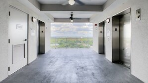 Ein Korridor mit zwei Aufzügen, einem Deckenventilator und Blick auf eine grüne Landschaft durch eine Glaswand.