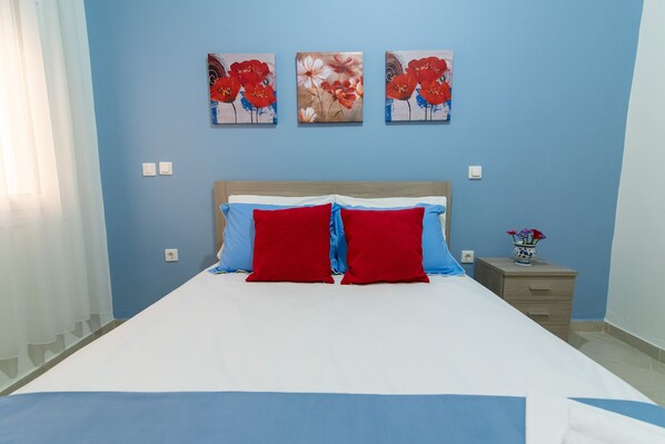 A bedroom with a bed, blue and red pillows, a wooden headboard, and three floral paintings on the wall.