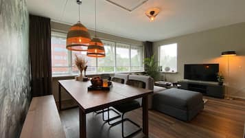 A modern living room with a wooden dining table, bench, sofa, and ottoman. There is a TV, a floor lamp, and a large window with curtains.