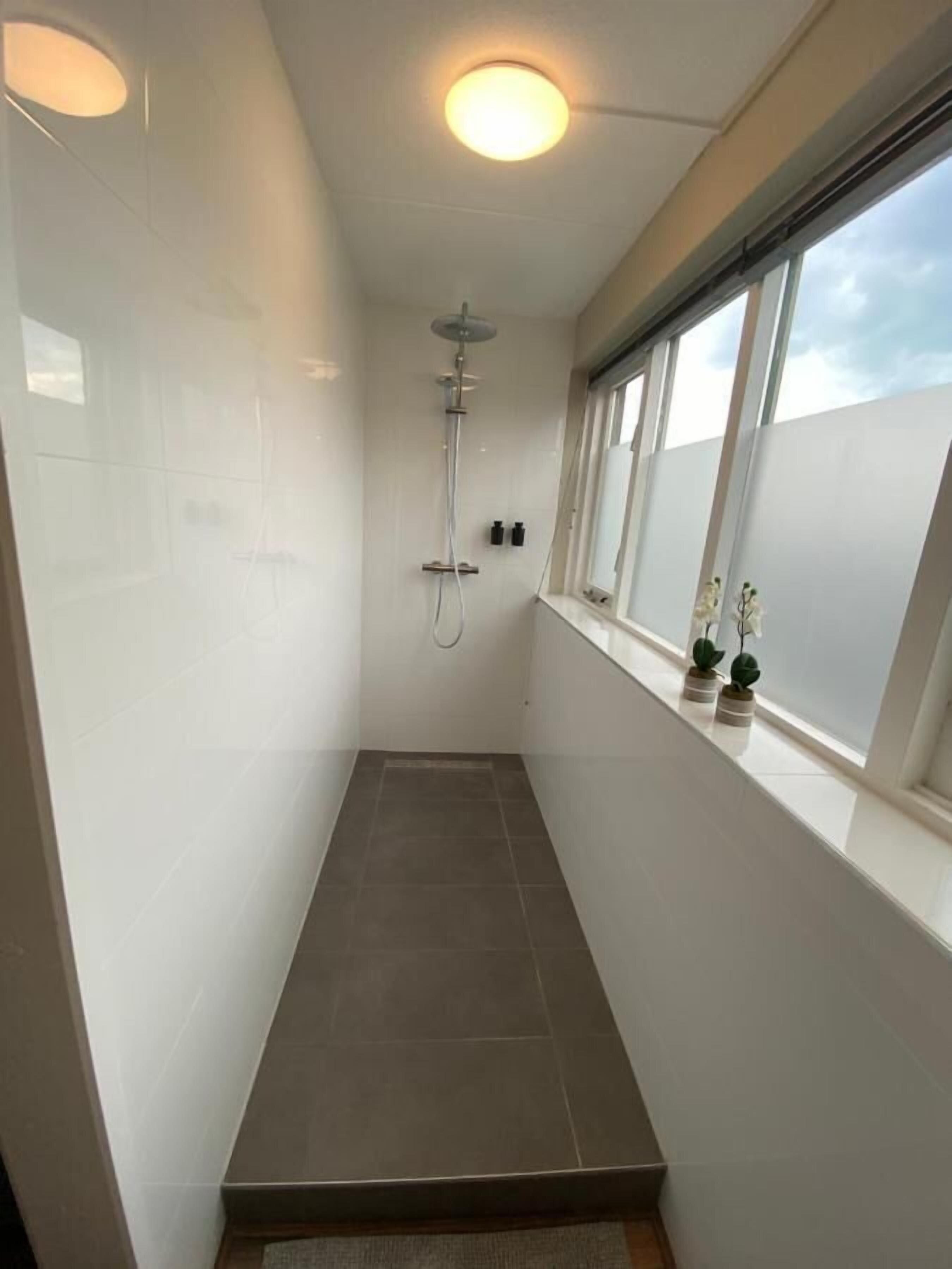 A modern bathroom with a shower, white tiles, a window with plants, and a ceiling light.