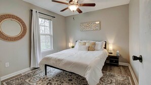 A bedroom with a bed, a ceiling fan, a window with curtains, a bedside table with a lamp, and a wall-mounted mirror.