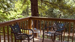 Una terraza de madera con dos sillas de metal y una pequeña mesa, rodeada de árboles.