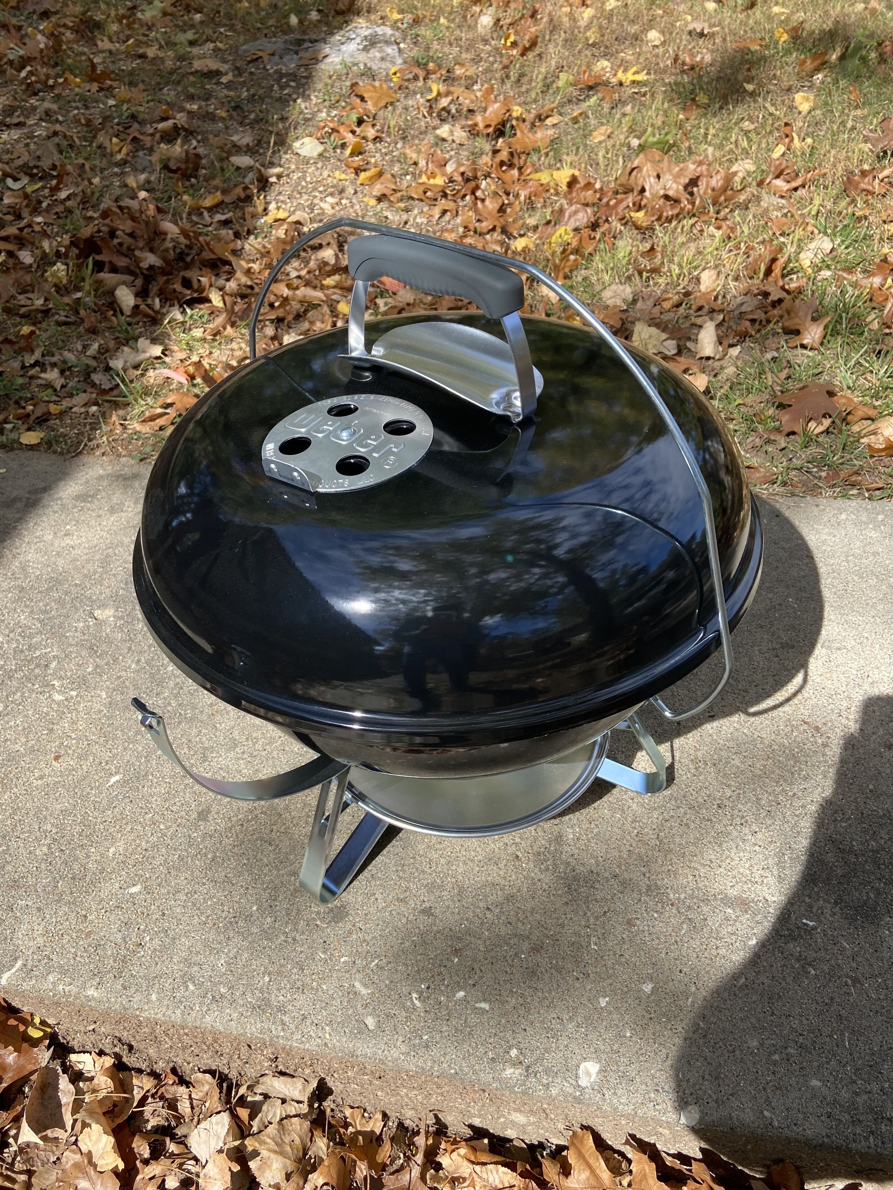 A black outdoor grill with a lid, placed on a concrete surface surrounded by dry leaves.
