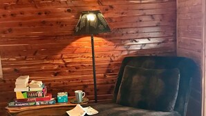 Una habitación acogedora con un sofá de terciopelo oscuro, suelo de madera, una lámpara, una mesita con libros y una taza a rayas, y una silla de mimbre con revistas.
