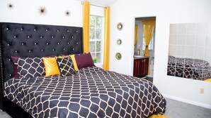 Une chambre à coucher avec une tête de lit capitonnée, une couvre-lit à motifs et une fenêtre ornée de rideaux jaunes.
