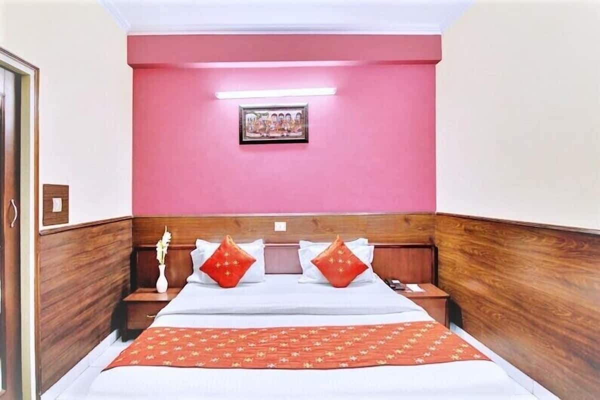 A hotel room with a wooden headboard, a bed with white bedding and red pillows, a red throw blanket, a bedside table with a vase, and a framed picture on the wall.