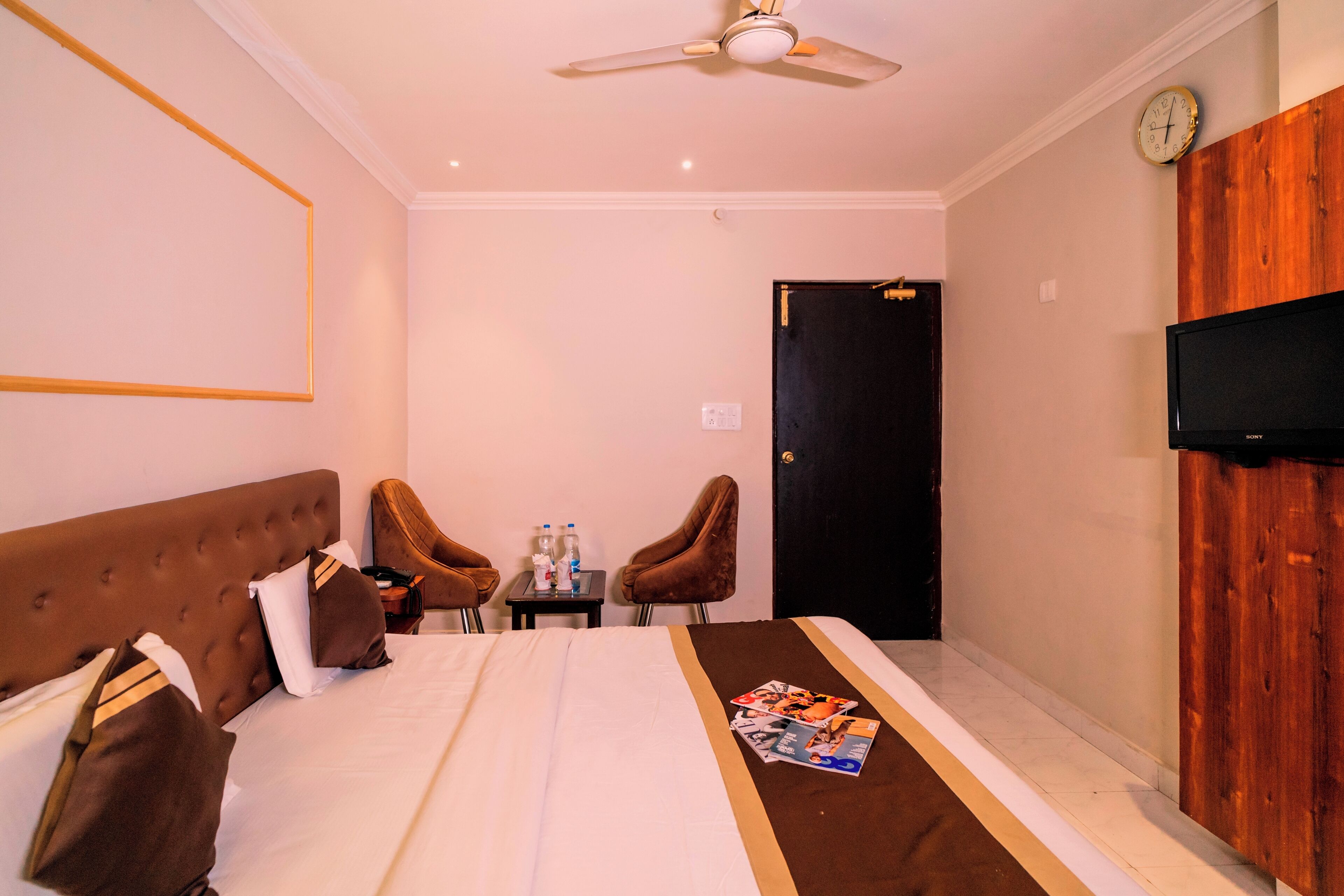 A hotel room with a bed, two chairs, a small table with bottles, a clock, and a TV on the wall.