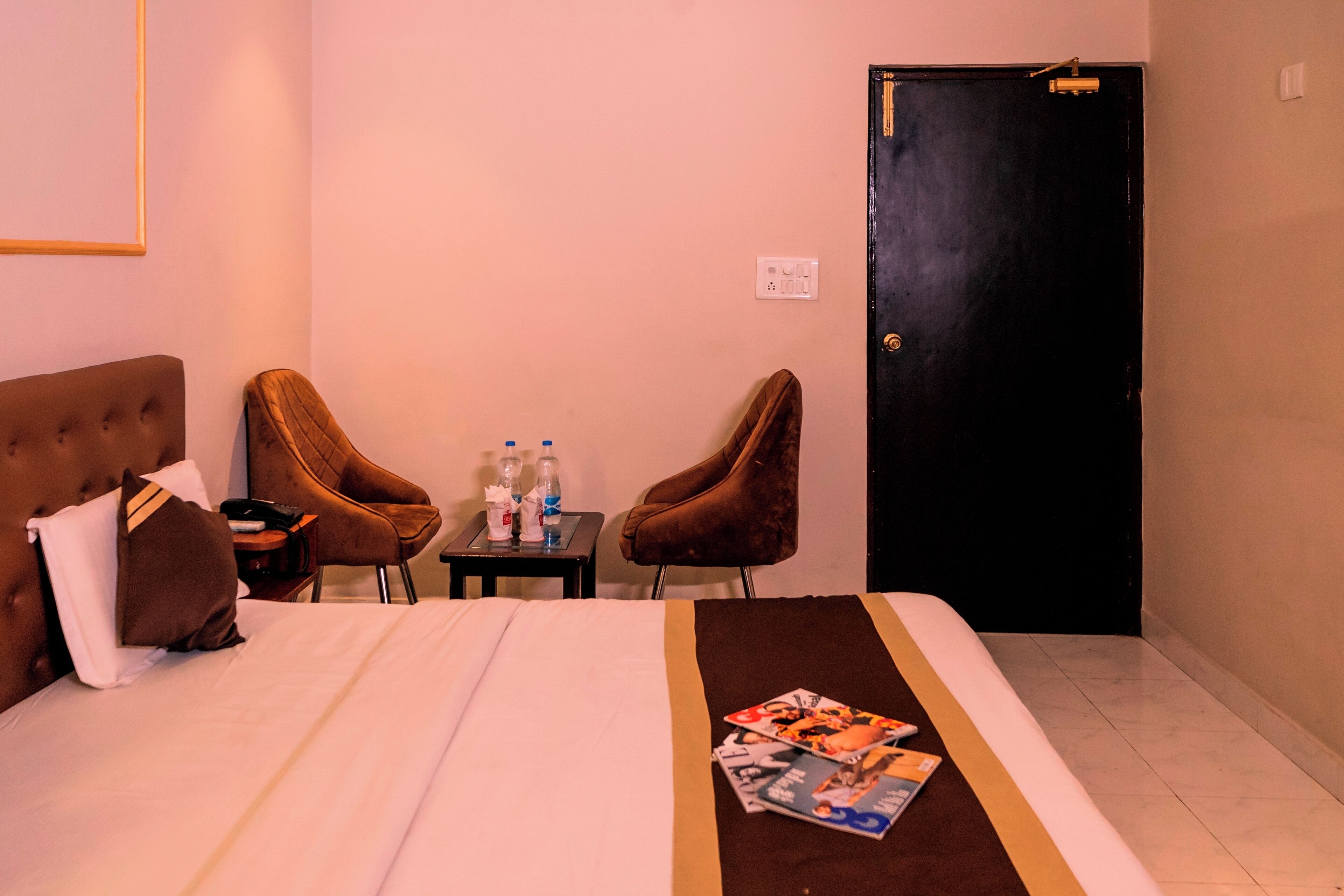 A hotel room with a bed, two armchairs, a small table with bottles, and a door.