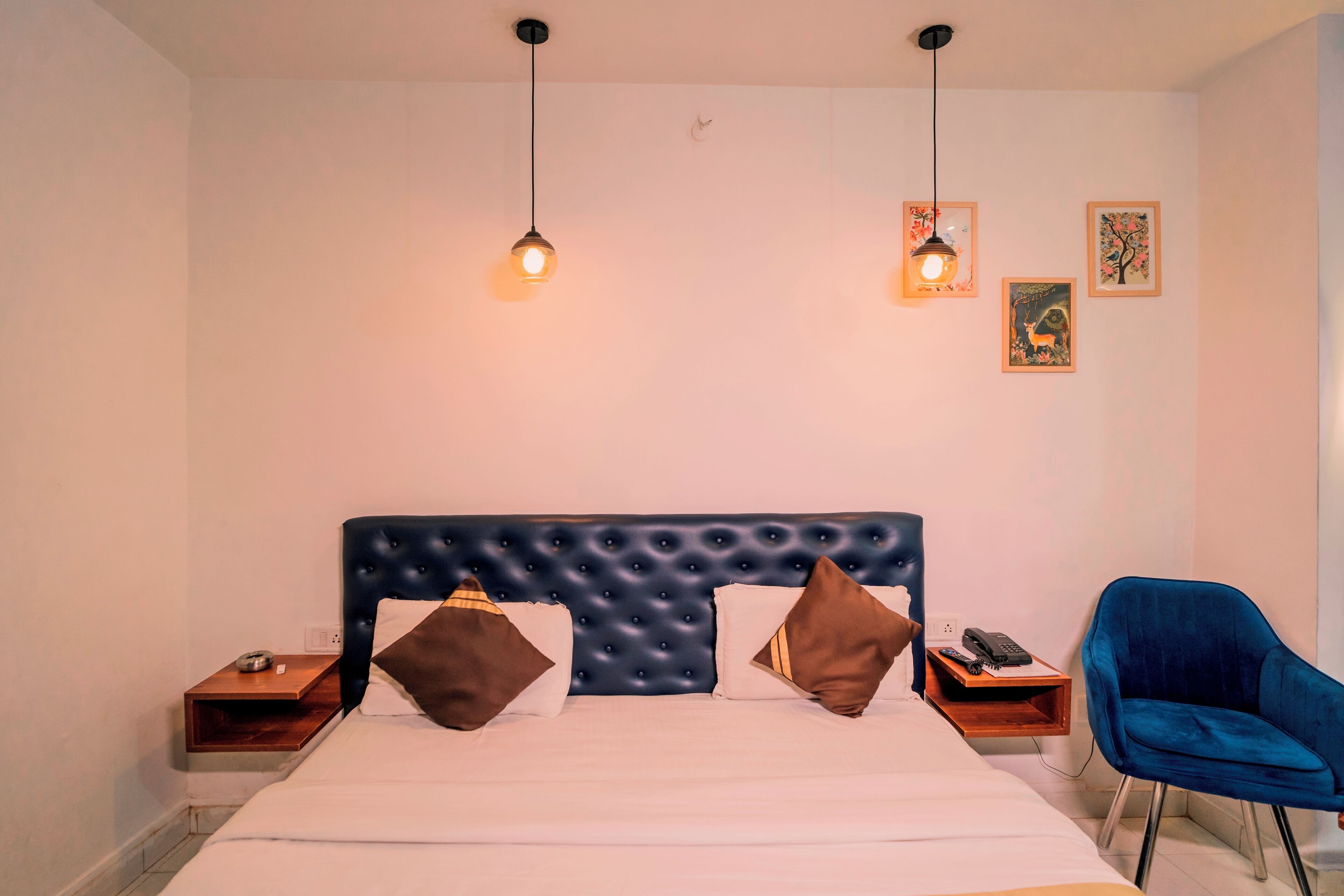 A neatly made bed with a tufted headboard, bedside tables with lamps, a blue chair, and framed pictures on the wall.