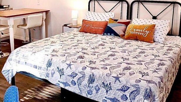 Ein Schlafzimmer mit einem Bett, einem Schreibtisch und einem Stuhl. An der Wand hängt ein Gemälde mit einer Strandansicht.