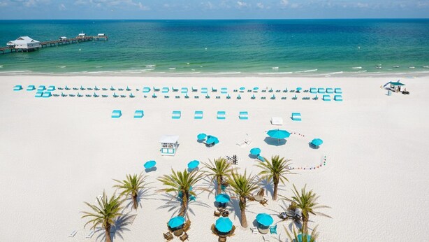 A beach with rows of blue umbrellas and lounge chairs, a pier in the background, and palm trees.