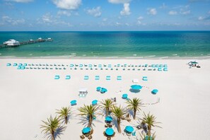 A beach with rows of blue umbrellas and lounge chairs, a pier in the background, and palm trees.