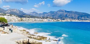 Un pueblo costero con edificios a lo largo de la orilla, una playa rocosa y montañas al fondo.